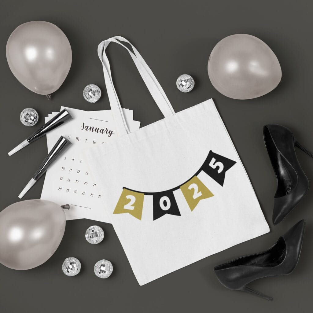 A white tote bag with a '2025' banner design in black and gold, surrounded by silver balloons and a New Year's calendar.