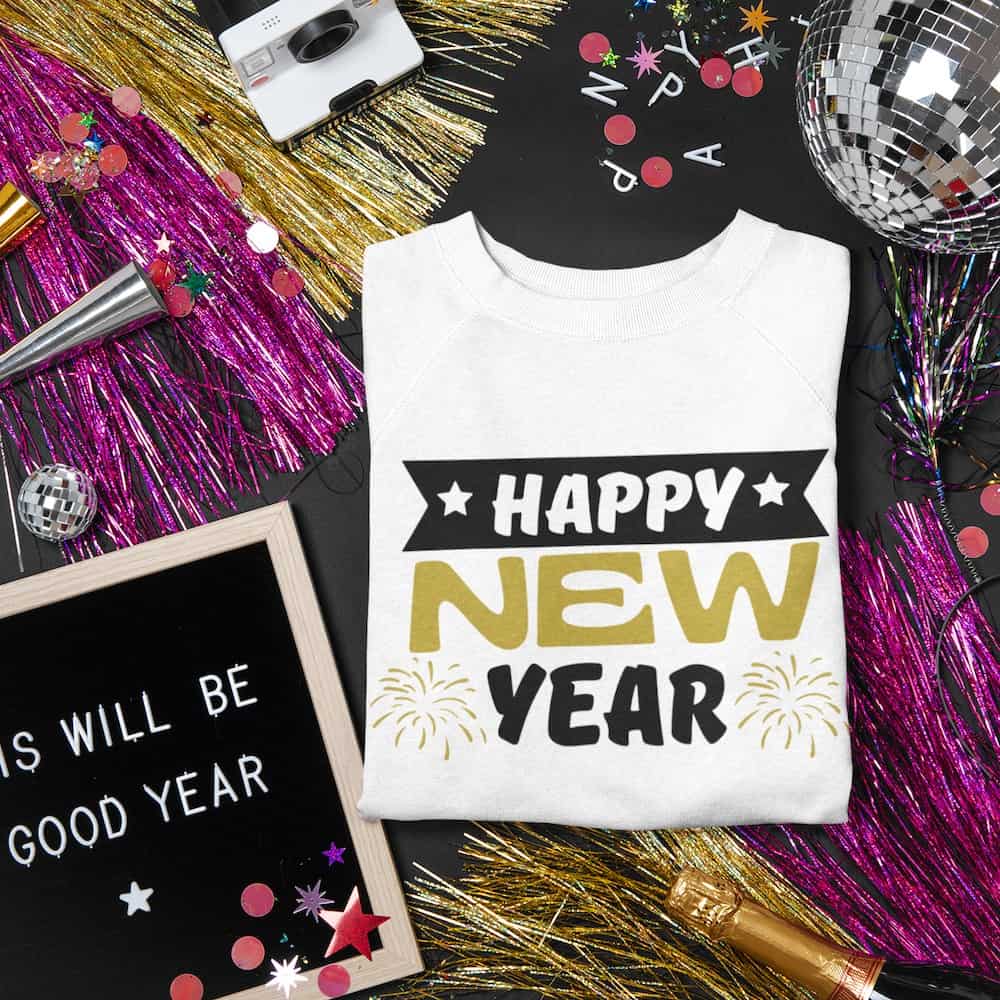 A folded white sweatshirt featuring a 'Happy New Year' design in black and gold with fireworks, styled against a festive backdrop with disco balls, tinsel, and celebratory decorations.