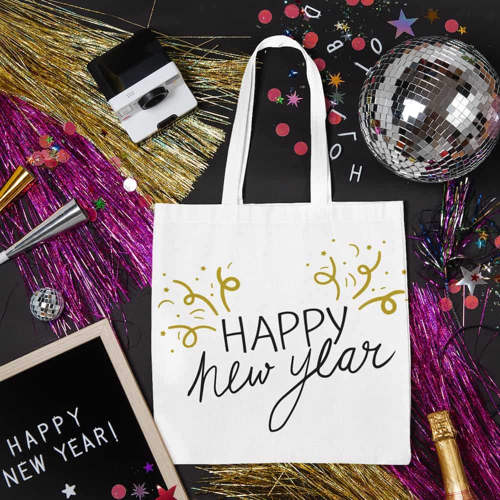 A white tote bag with 'Happy New Year' handwritten text and gold confetti accents, styled with disco balls and festive party decorations.