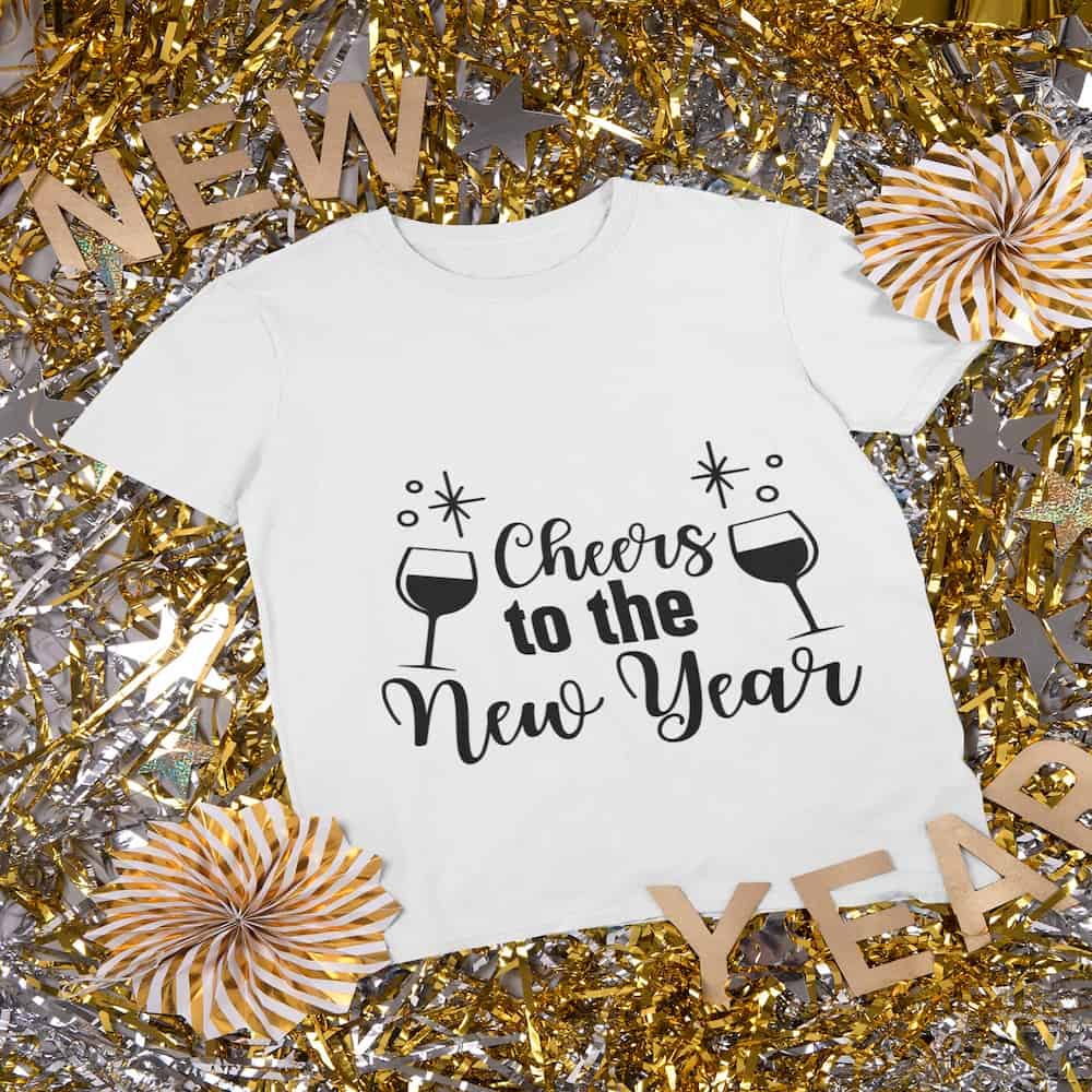 A white t-shirt displaying 'Cheers to the New Year' text with wine glasses and star accents, surrounded by gold and silver party tinsel.