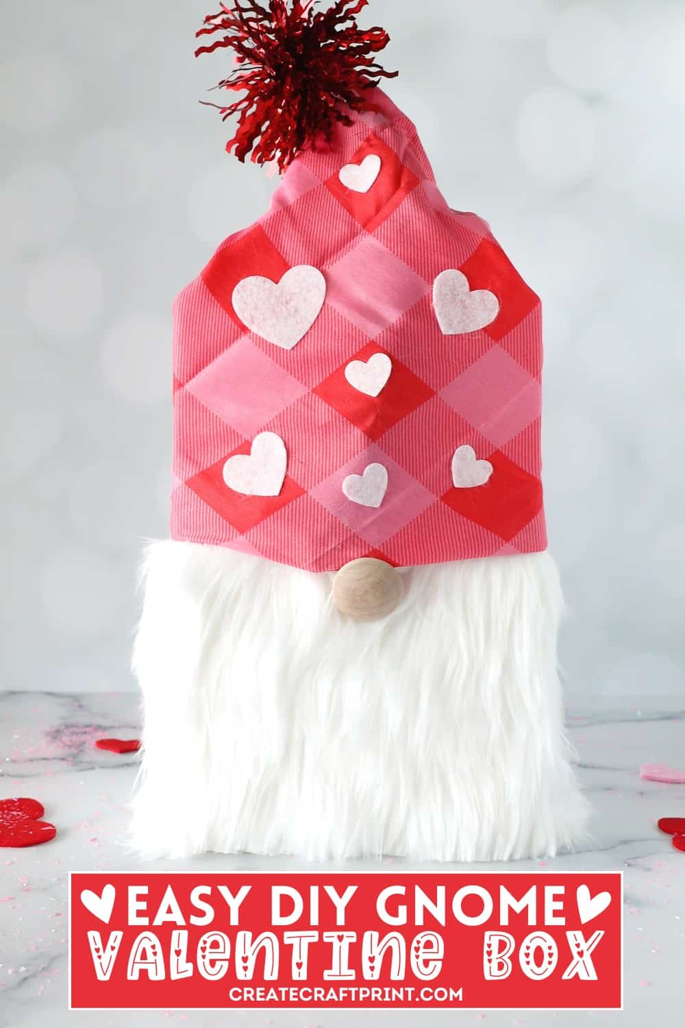 A DIY Gnome Valentine Box featuring a pink and red checkered hat with felt hearts, a fluffy white beard made of craft fur, and a wooden knob nose. The box is topped with a red pom-pom, set against a light, festive background with scattered heart decorations.