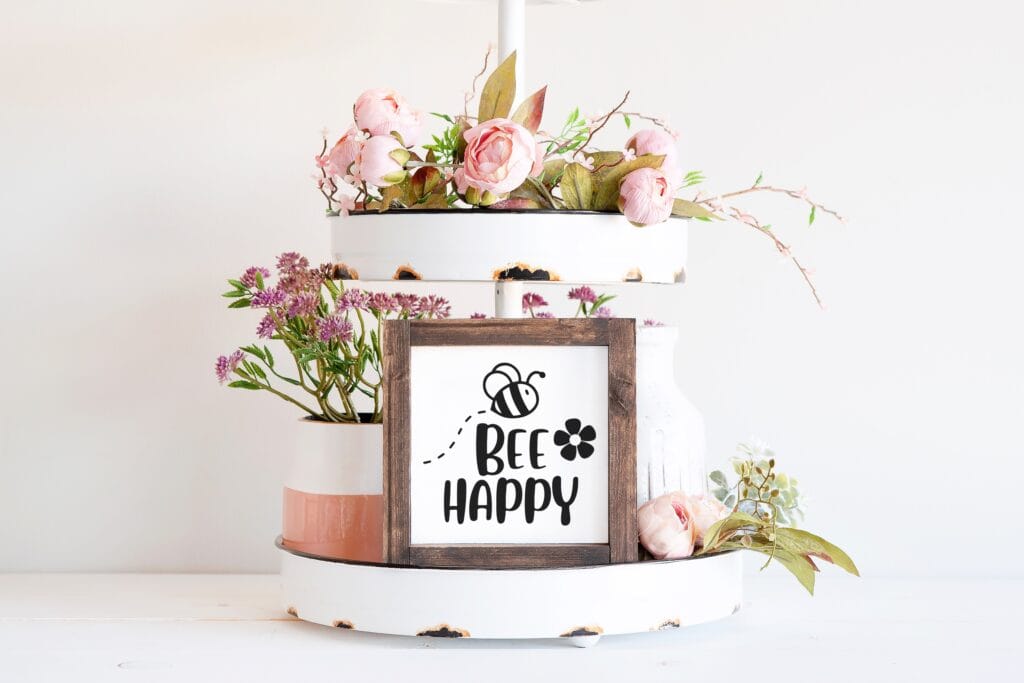 Rustic tiered tray display featuring a wooden farmhouse-style sign with 'Bee Happy' SVG design, surrounded by pink flowers and greenery, perfect for spring Cricut crafts.