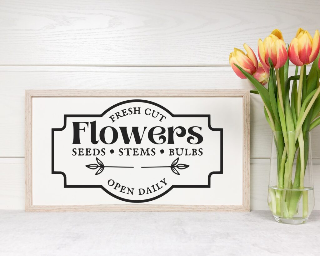 Farmhouse-style 'Fresh Cut Flowers' SVG sign in a light wood frame, styled with a vase of tulips on a white rustic surface, ideal for spring Cricut home decor projects.