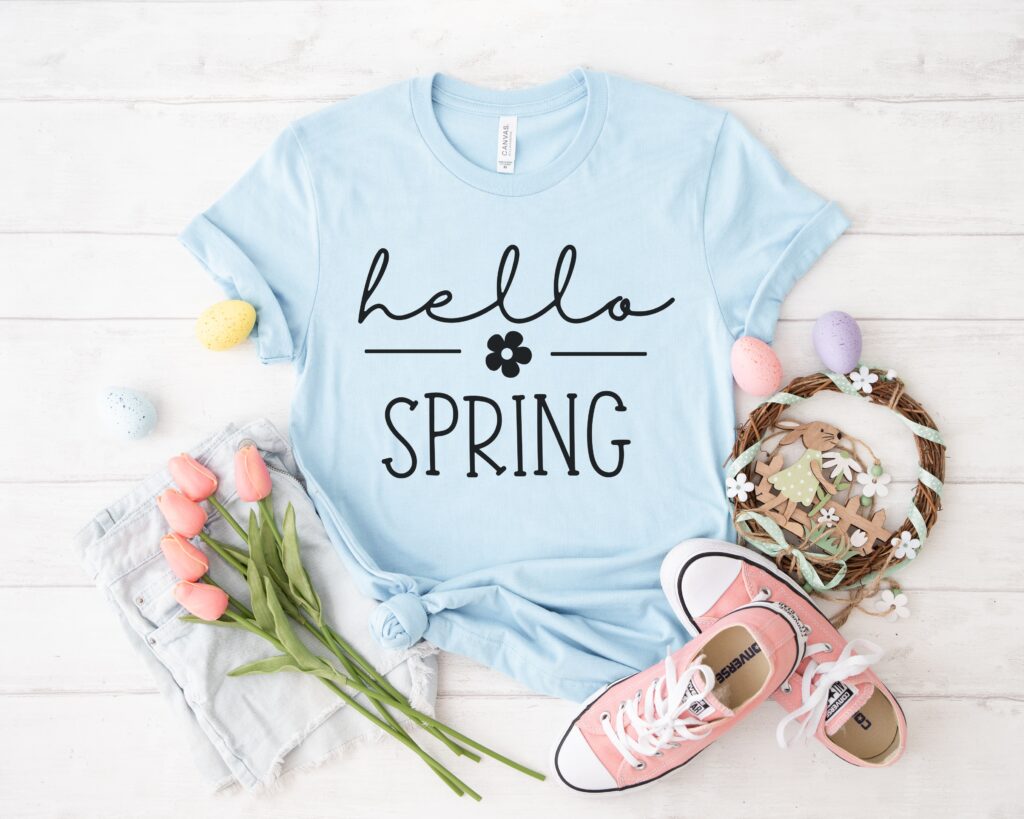 Flat lay of a light blue t-shirt featuring 'Hello Spring' design in black, styled with pastel Easter eggs, tulips, pink sneakers, and denim shorts, ideal for spring Cricut apparel projects.