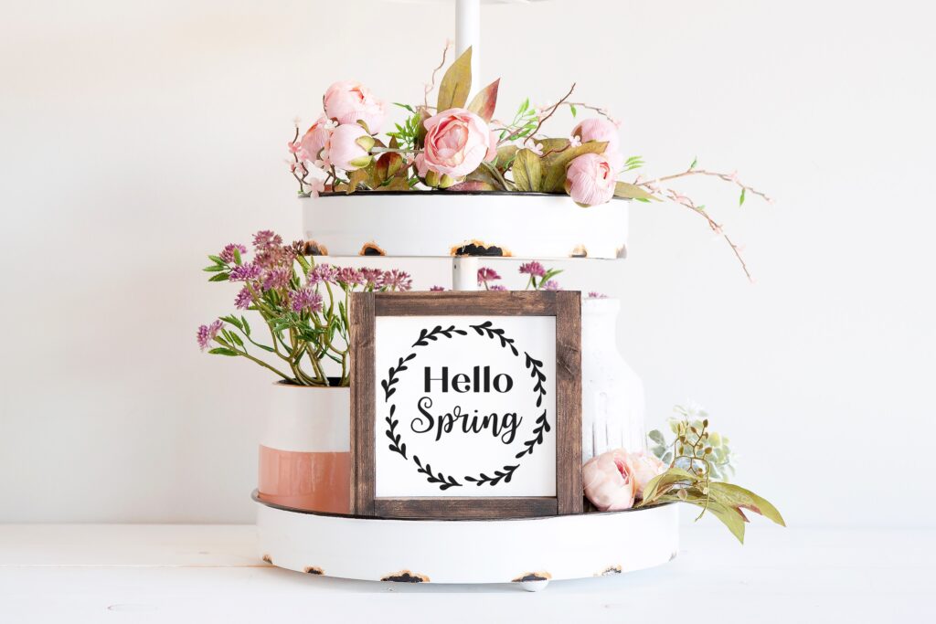 Rustic farmhouse-style tiered tray display featuring a framed 'Hello Spring' wreath sign, styled with pink flowers and vintage decor, perfect for spring home crafts.