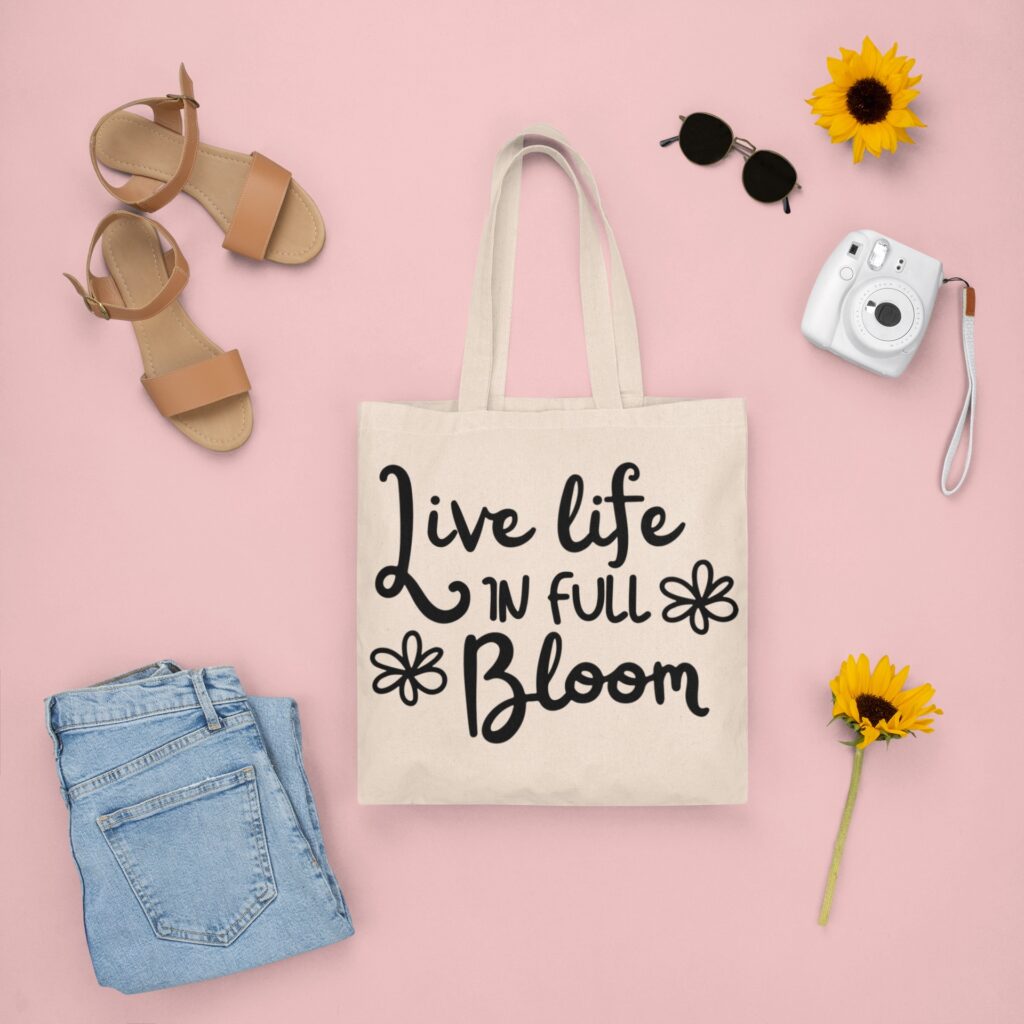 Spring-themed tote bag featuring 'Live Life in Full Bloom' SVG design in black, styled with denim jeans, tan sandals, sunflowers, sunglasses, and a vintage camera on a pastel pink background.