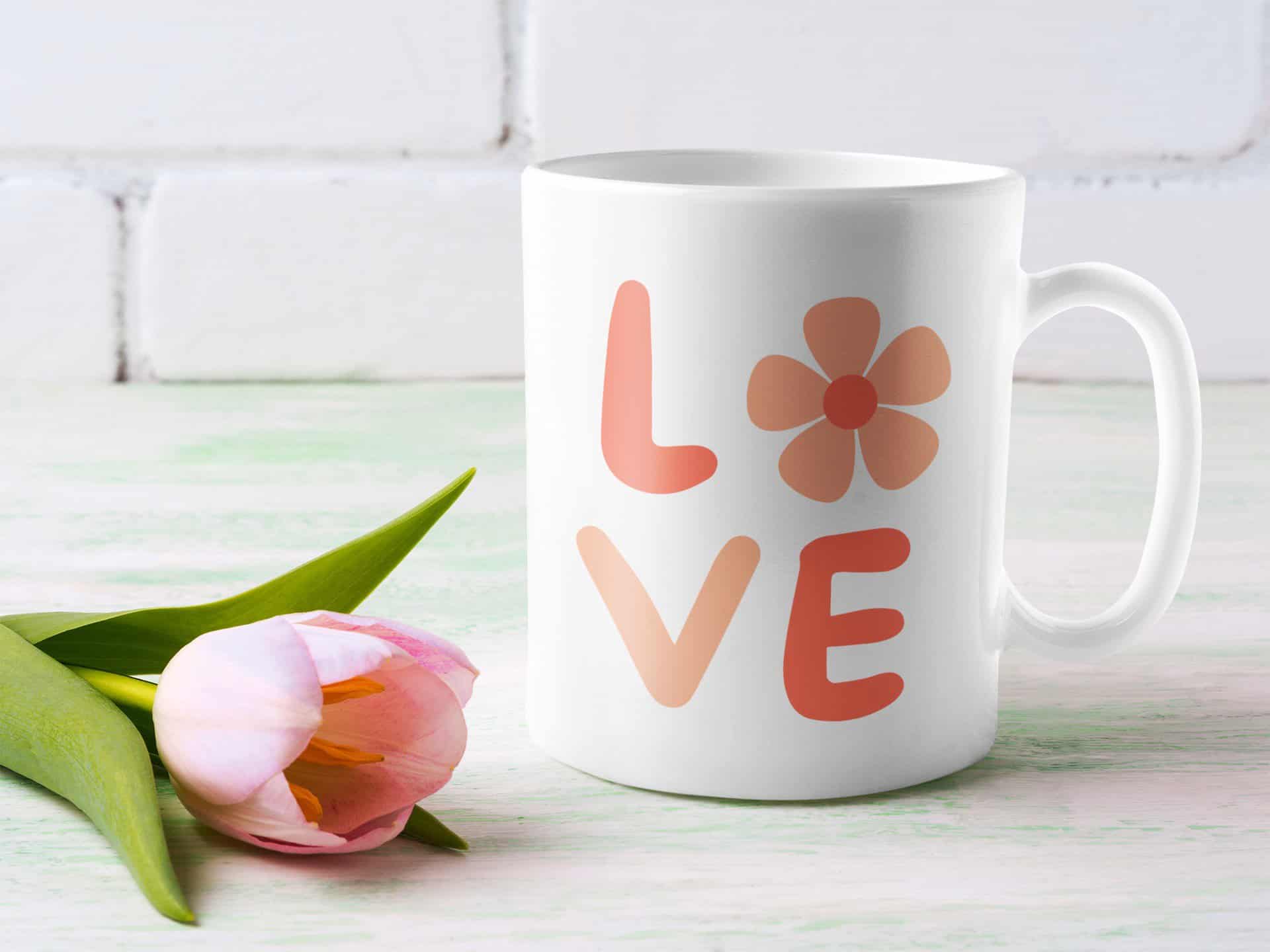 White ceramic mug with 'LOVE' floral SVG design in warm peach and pink tones, styled with a fresh pink tulip on a pastel wooden background, ideal for DIY spring Cricut projects.