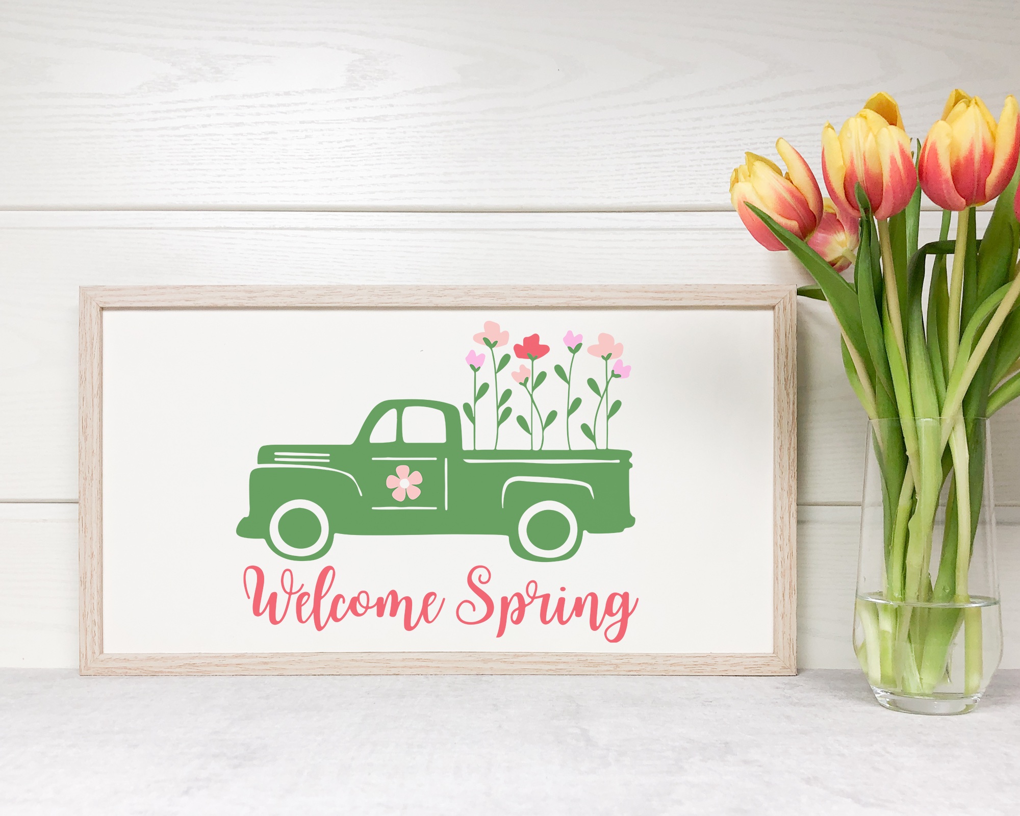 Rustic farmhouse-style framed sign featuring a vintage green truck with pink flowers and 'Welcome Spring' design, styled with a vase of tulips, ideal for home decor crafts.