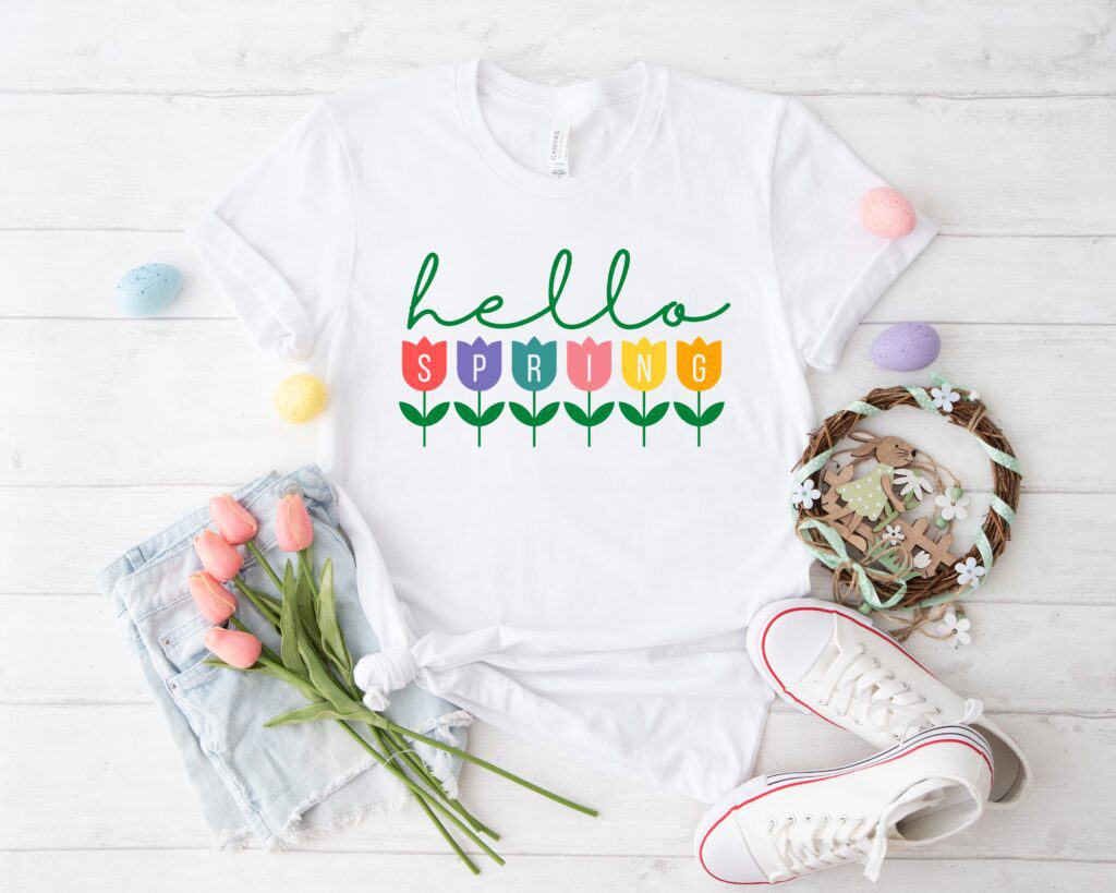 Flat lay of a white t-shirt with a colorful 'Hello Spring' tulip SVG design, styled with Easter decor, pink sneakers, and tulips, perfect for Cricut and Silhouette DIY projects.