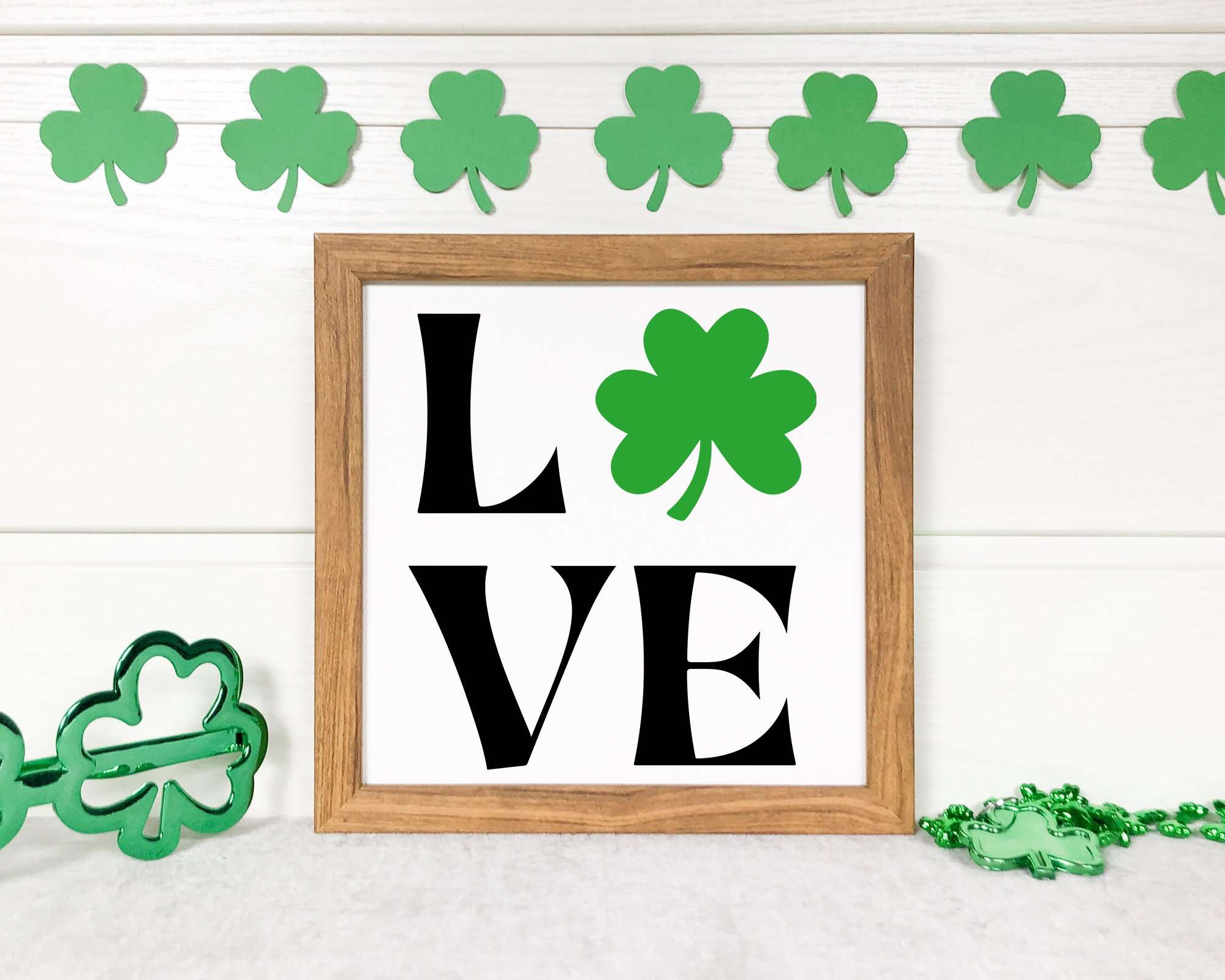 Framed 'LOVE' sign with black typography and a green clover, surrounded by festive shamrock garland. Ideal for DIY St. Patrick’s Day home décor and Cricut crafting.