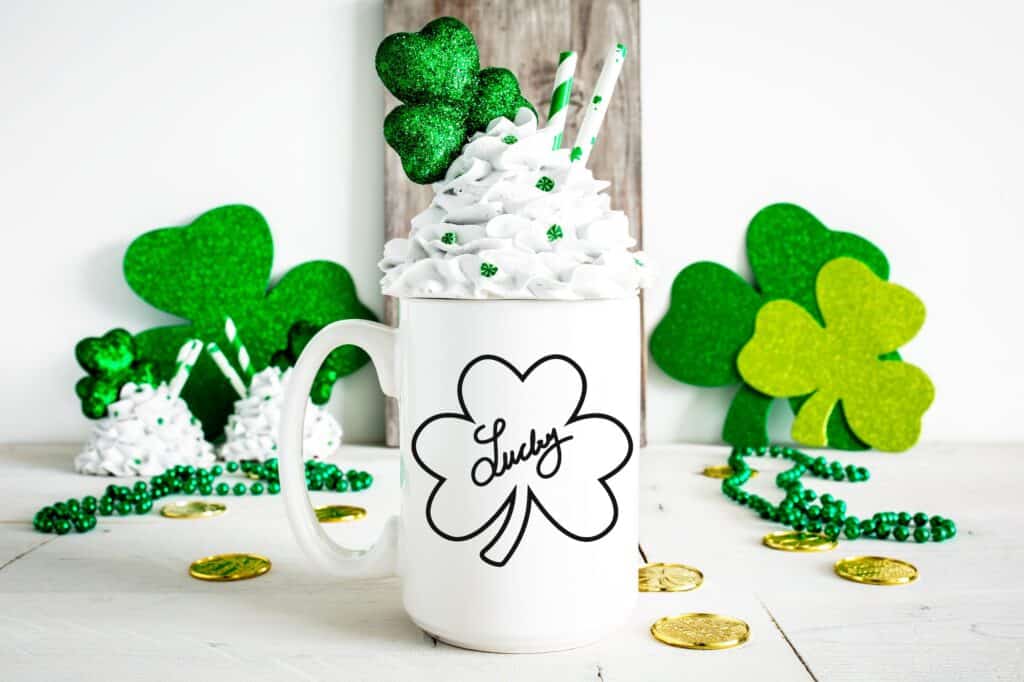 White mug with a 'Lucky' shamrock outline SVG design, topped with whipped cream and surrounded by glittery green shamrocks and gold coins.