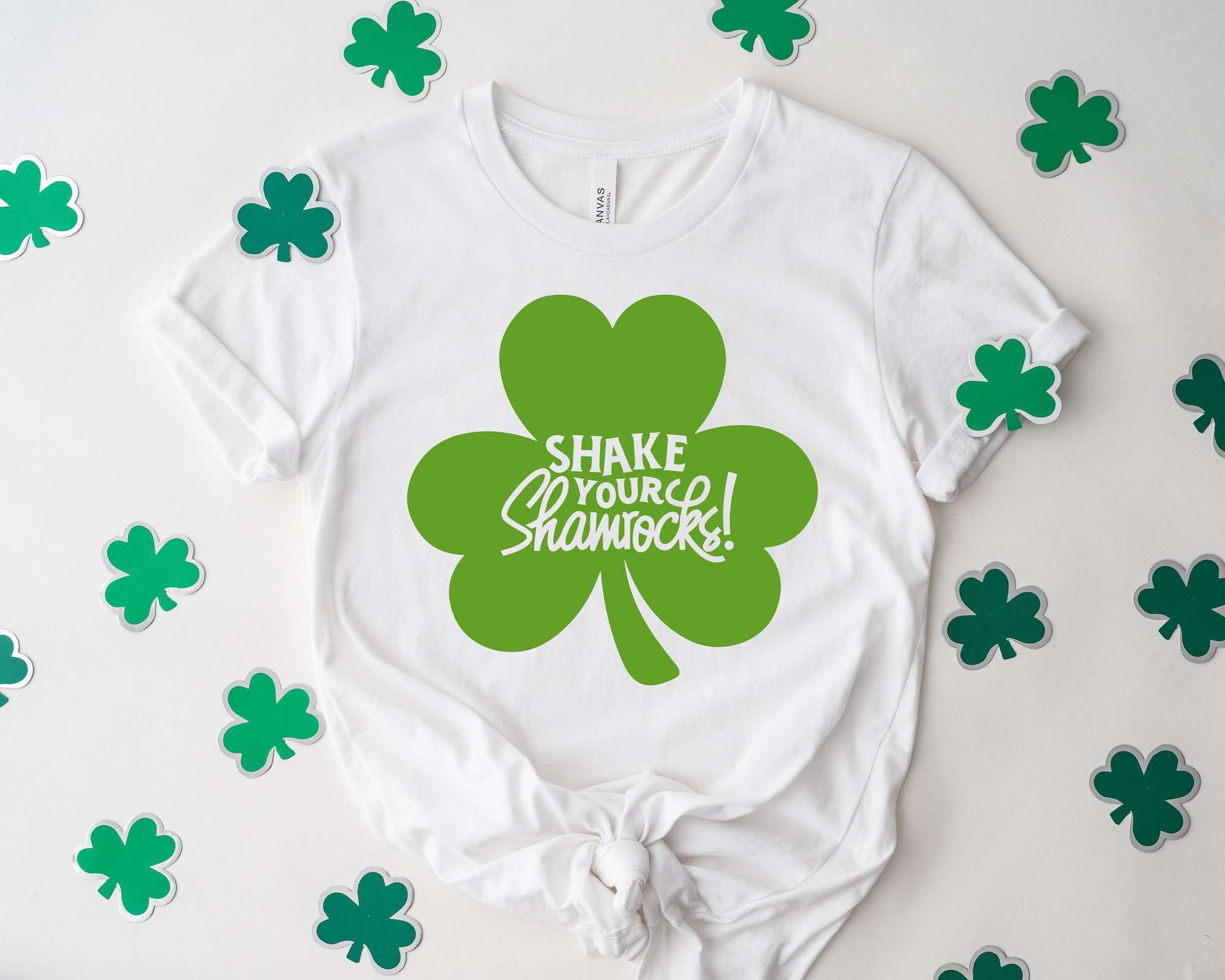 White t-shirt with a bold green 'Shake Your Shamrocks' design, surrounded by scattered shamrock cutouts. 