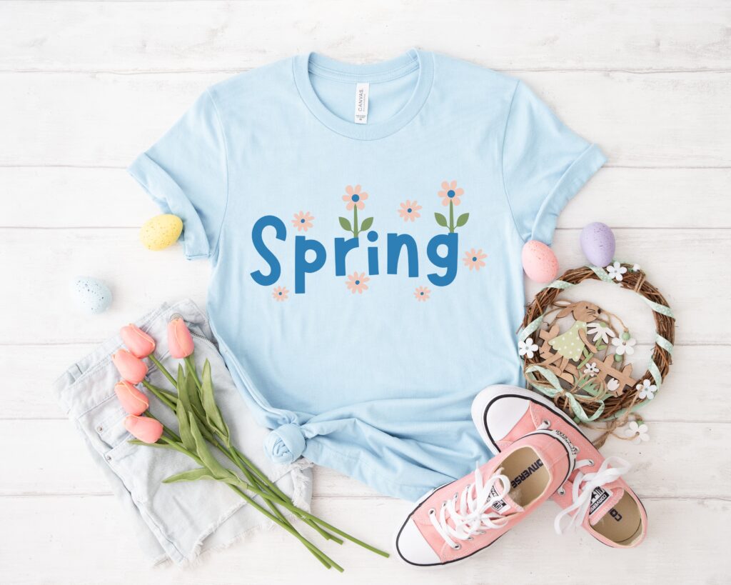 Flat lay of a light blue t-shirt featuring 'Spring' design with floral accents, styled with pastel Easter eggs, pink tulips, sneakers, and denim shorts, perfect for spring Cricut apparel crafts.