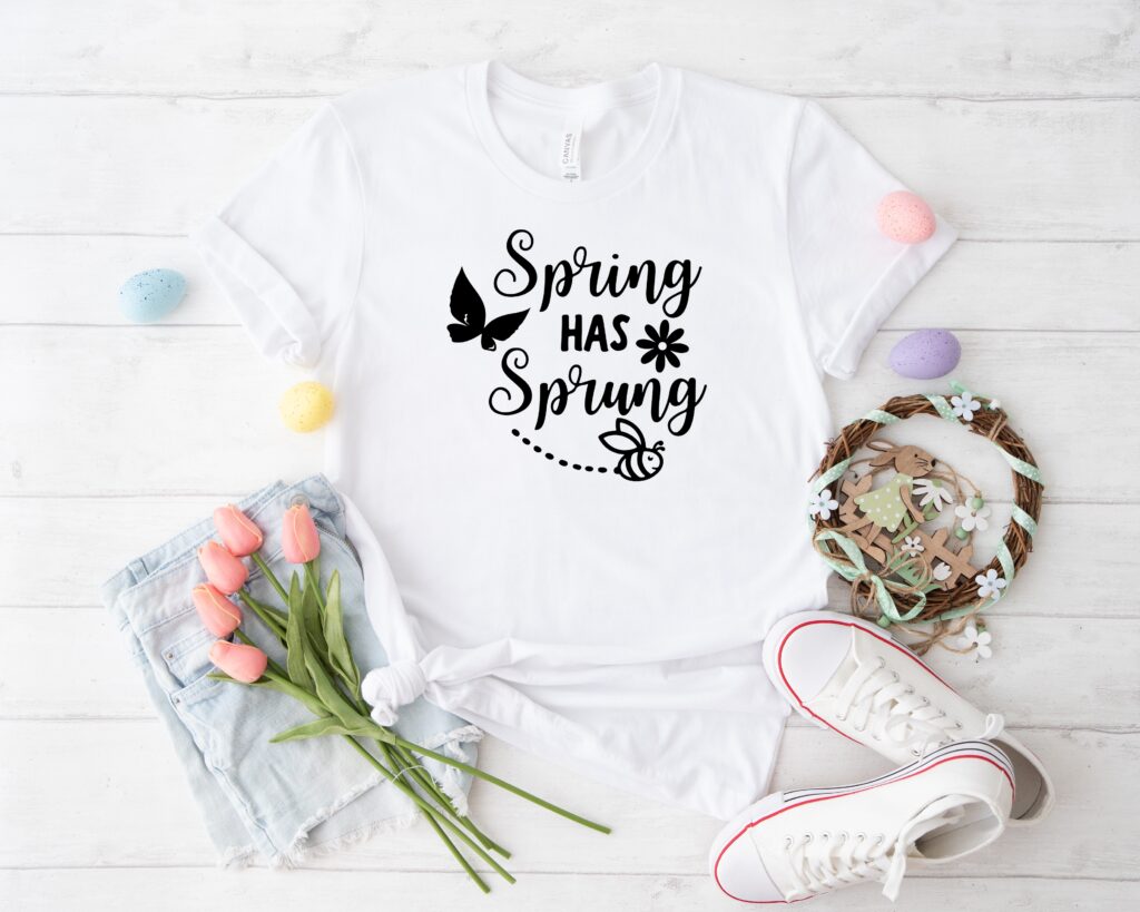 Flat lay of a white t-shirt featuring 'Spring Has Sprung' SVG design with a butterfly and bee, surrounded by Easter decor, perfect for seasonal DIY Cricut and Silhouette crafts.